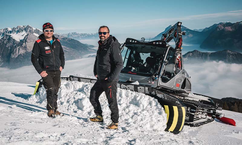 Two people standing in front of a Prinoth snowgroomer equipped with Leica iCON alpine snow management system. 