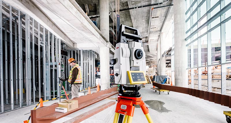 Construction worker with orange vest and safety hat in building construction site, looks in an Leica iCON CC80 field controller connected to a Leica iCON iCR70 construction robotic total station.