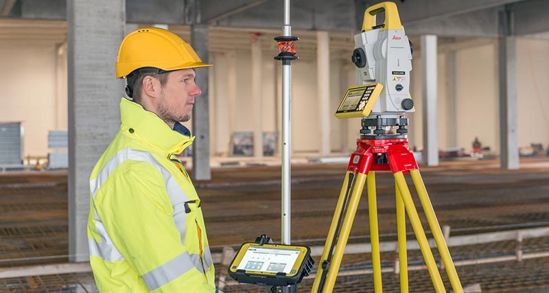 Construction worked in yellow west and hard hat holds a prism pole with the Leica iCON CC80 field controller with iCON build software and is connected to a Leica iCON iCR80 robotic total station.