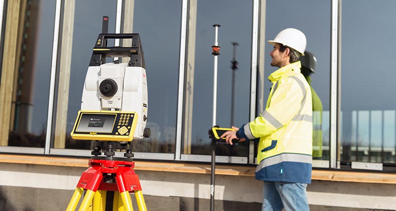 Építőmunkás sárga mellényben és kemény sisakban egy prizmarudat tart a Leica iCON CC80 terepi kontrollerrel és az iCON build szoftverrel, és csatlakozik egy Leica iCON iCR80 robot mérőállomáshoz, stabil célkövetéssel kitűzés közben.