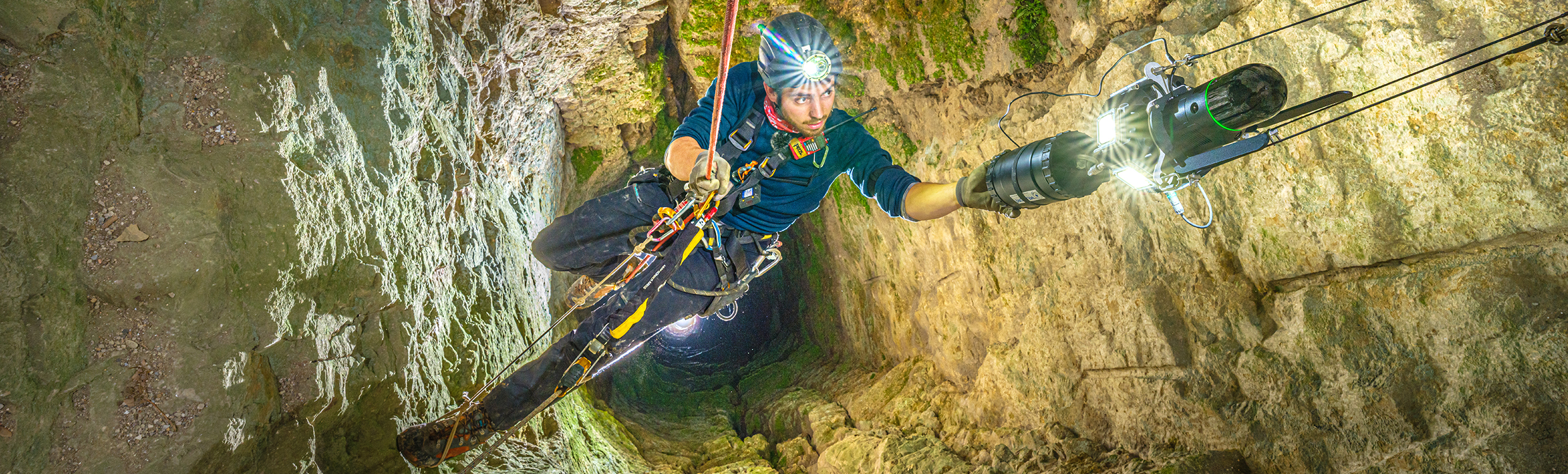 A man supported by rappelling gear scans inside a cave with a laser scanner